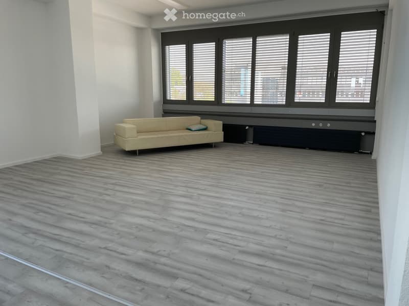 Empty room with white walls, gray wooden floor, beige couch, large windows with blinds, and a view of the outside.
