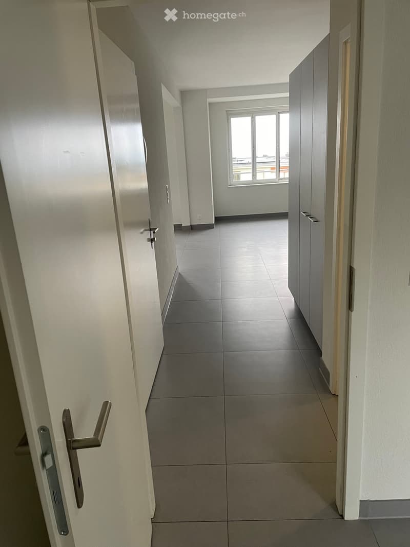 Empty hallway with white walls, gray tiles, and a window with a view outside.
