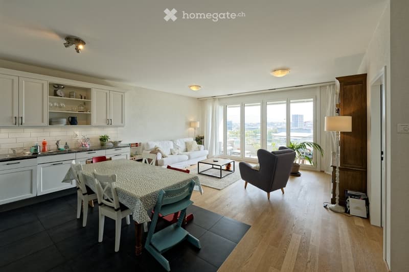 Open plan living room and dining area with large windows, white kitchen, dining table, couch, armchair, coffee table, floor lamp, rug, potted plant