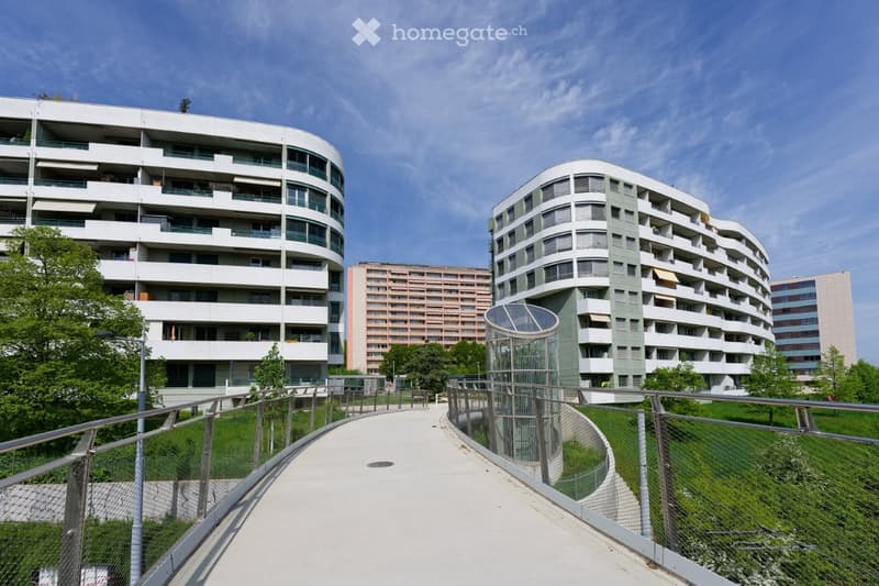 Two modern apartment buildings, white and green exterior, multiple floors, balconies, surrounded by greenery and a pathway