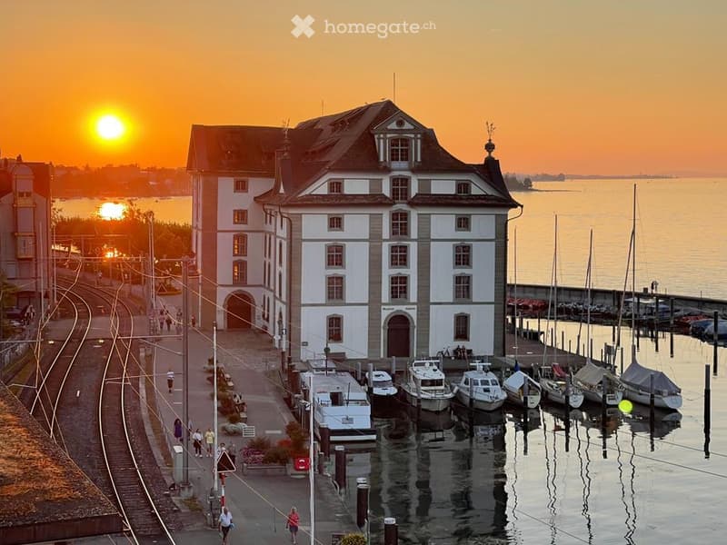A large white building with a brown roof, situated on the waterfront with a dock for boats, surrounded by a scenic sunset view.