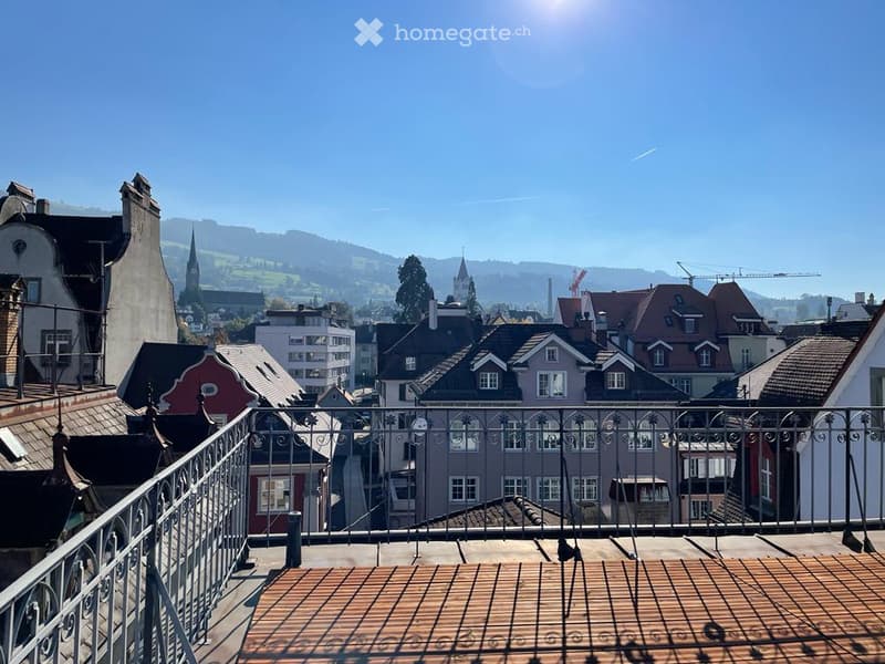 Aerial view of a city with multiple buildings, a balcony with a metal railing, and a view of the mountains in the distance.