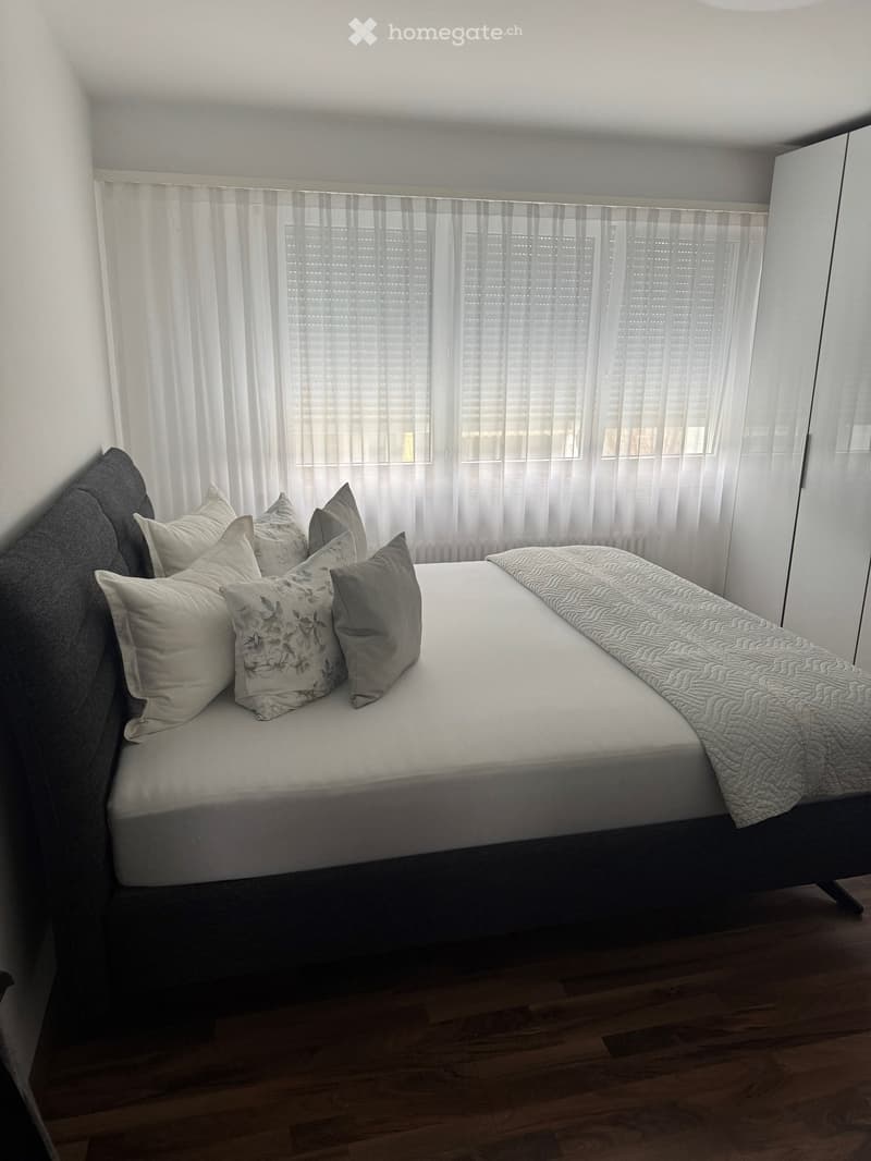 Bedroom with a bed, pillows, and a blanket, white walls, wooden floor, white curtains, and a white wardrobe