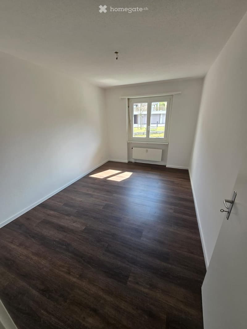 Empty room with white walls, wooden floor, window with white curtains, white radiator