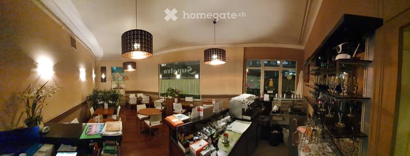 A restaurant with a wooden floor, beige walls, a counter, tables, chairs, lamps, plants, and a glass window with a sign