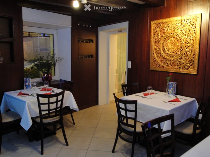 Furnished Room. A restaurant with wooden walls and tiled floor. There are two tables with white tablecloths, each set with wine glasses, napkins, and utensils. A flower vase is on one of the tables. There is a large decorative wall art on the right wall.