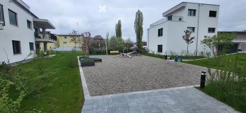 A gravel path leads to a small playground with a swing and a bench. The garden is surrounded by grass and has a small tree and a planter.
