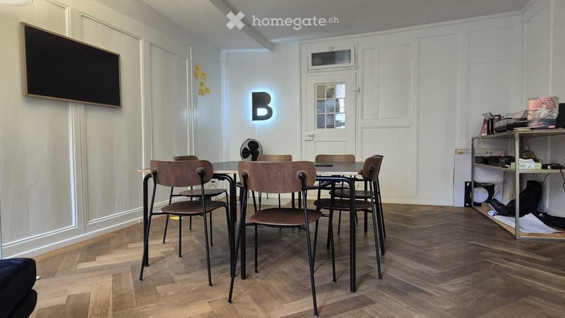 Wooden floor, white walls, round table, 6 chairs, blackboard, fan, shelves, door