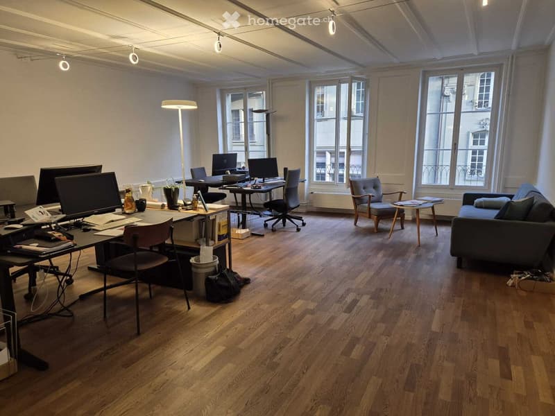 Office space with desks, chairs, computers, and a couch. Wooden floors, white walls, and large windows.