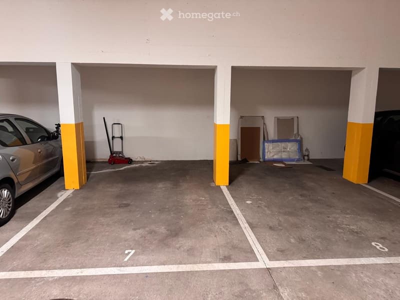 Underground parking area with two cars parked, yellow and white pillars, empty parking spaces, some storage boxes and a red trolley