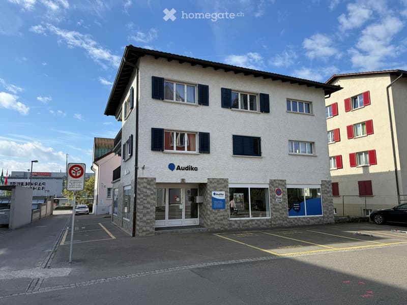 2 story building, white exterior, blue shutters, Audika logo, parking lot in front