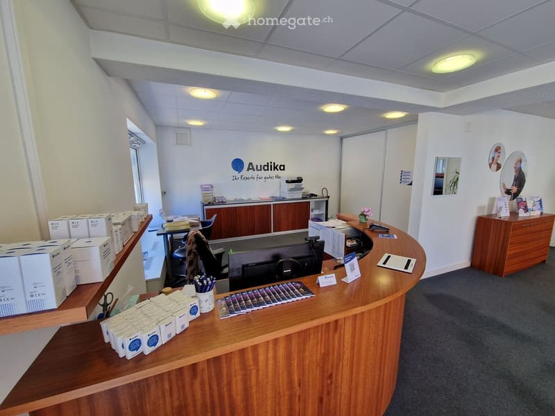 Reception area with a wooden desk, computer, office supplies, and a wooden cabinet with a flower vase. A wall with a logo and a mirror. A wooden shelf with boxes and scissors.