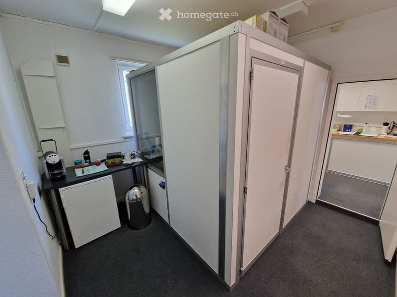 A small room with a white wall, a white door, a small table with a coffee machine, a trash bin, and a small refrigerator. There is a glass door with a window and a small shelf with some items on top. There is a small cabinet with a white door and a small