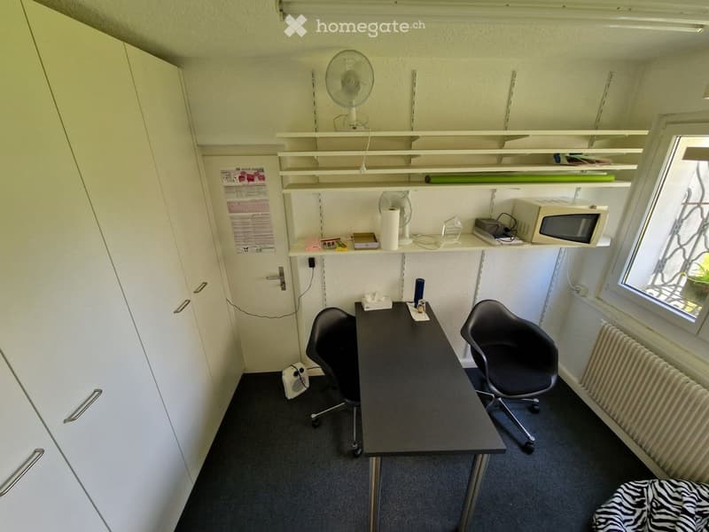 small room with desk, two chairs, shelves, radiator, cabinets, microwave, fan