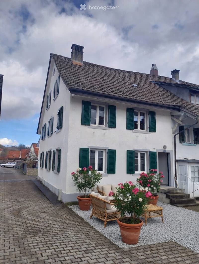 2 story house, white exterior, green shutters, brick driveway, front garden with plants and outdoor seating