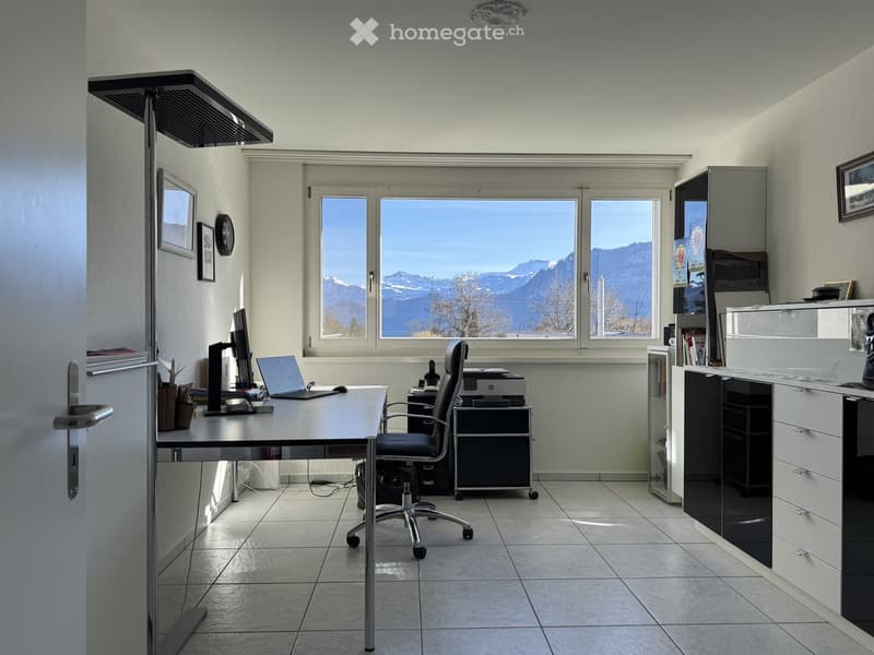 An office space with desk, office chair, laptop, monitor, printer, and mountain view through a large window.