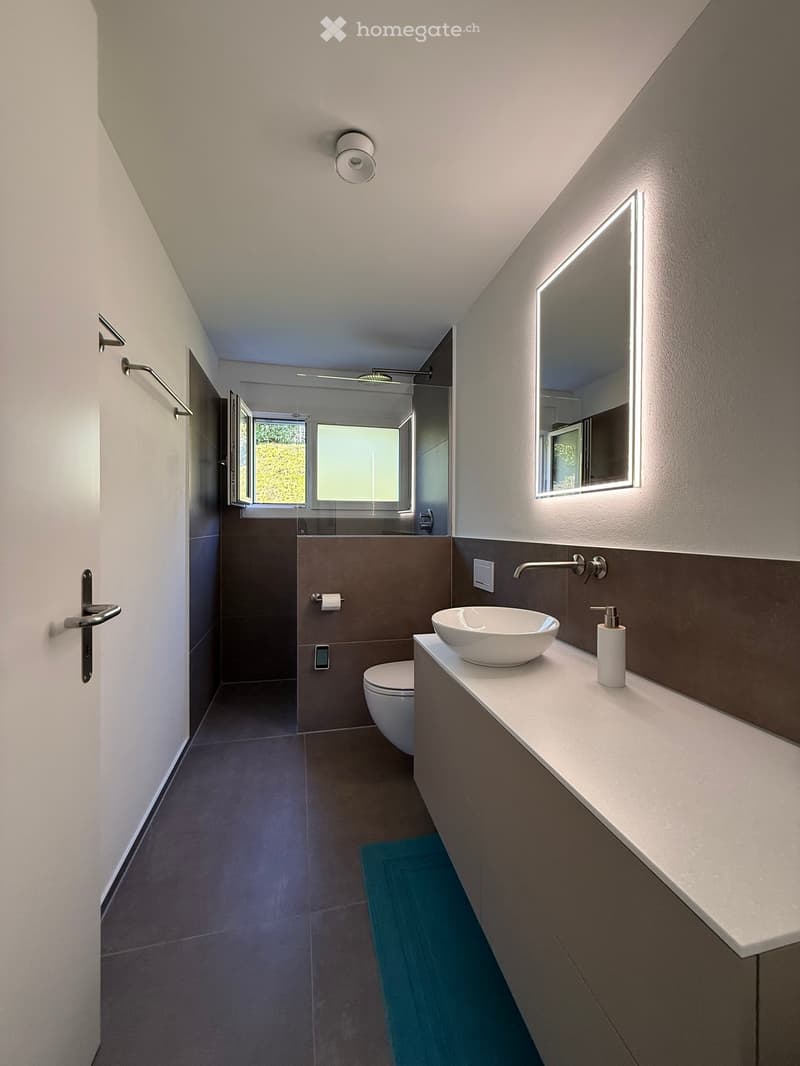 modern bathroom with tiled floors, white vanity, white sink, mirror with lighting, separate toilet, glass shower door