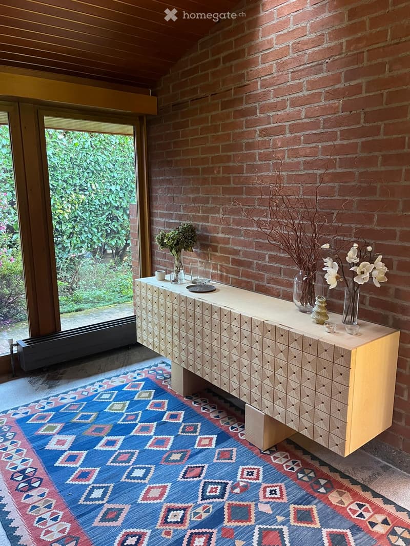 A room with a wooden console table with geometric design, brick wall, glass door with a white blind, a rug, and greenery.