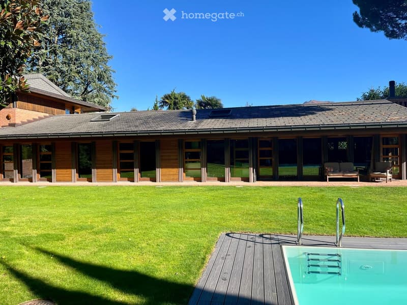 House with wooden facade, large windows, swimming pool, wooden deck, well-maintained lawn, surrounded by trees.