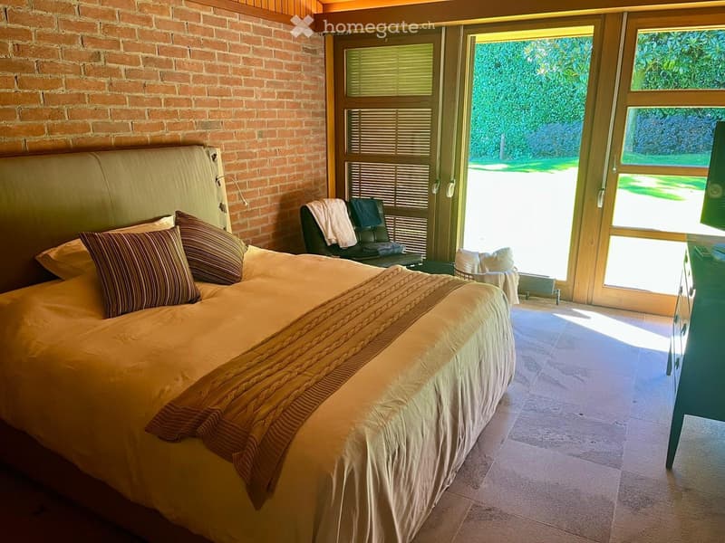 bedroom with brick wall, double bed, windows, green outdoor view, couch, cabinet
