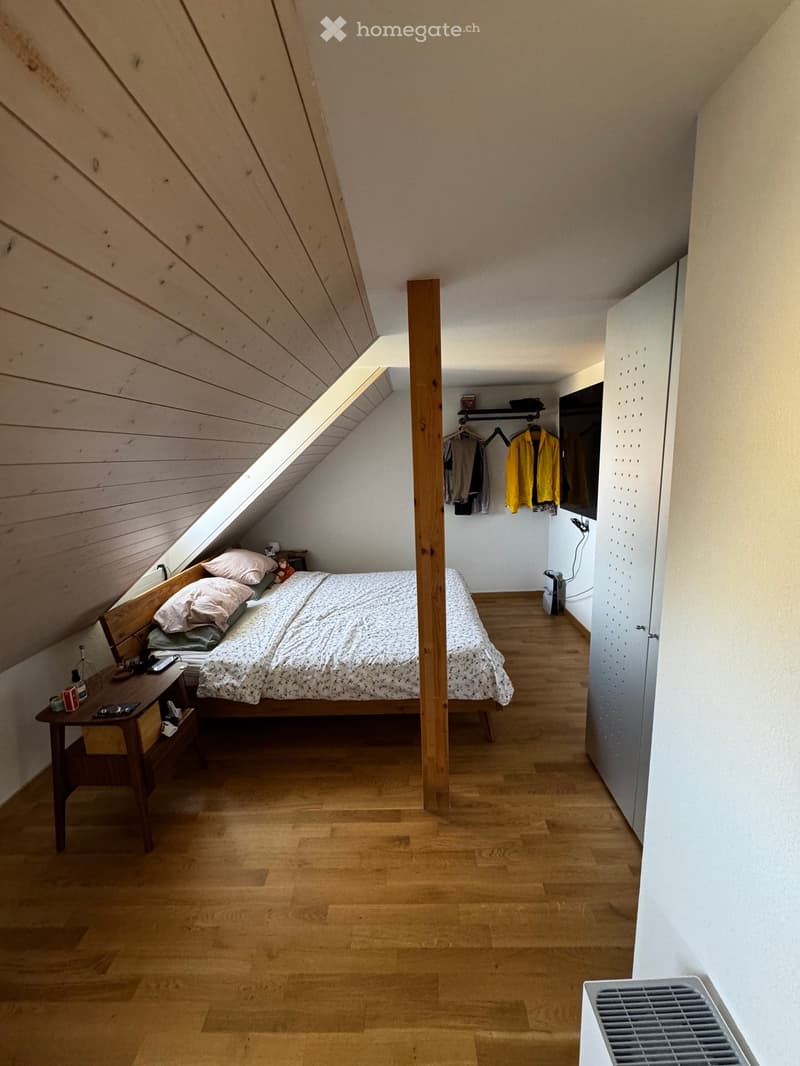 Bedroom with wooden bed, wooden nightstand, wooden ceiling, wooden floor, white walls, clothes hanging on the wall, TV mounted on the wall, and a small shelf with items on it.
