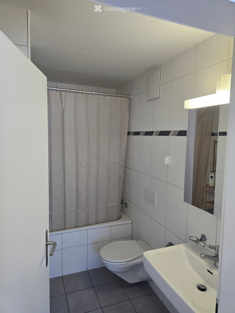 Bathroom with white tiles, bathtub, shower curtain, toilet, sink, mirror, and a small shelf