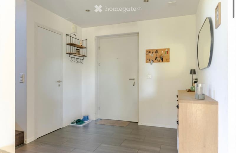 Empty hallway with white walls, wooden floor, two white doors, a shelf with hooks, a mirror, a lamp, and a wooden cabinet with drawers.