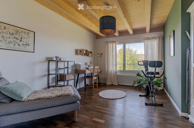 A room with a couch, desk, chair, exercise bike, shelves, and a window with curtains.