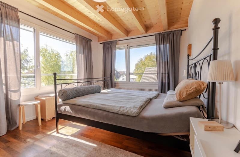 Bedroom with wooden floor, large windows, bed, nightstands, lamp, rug, wooden stool, wooden headboard, pillows, blanket