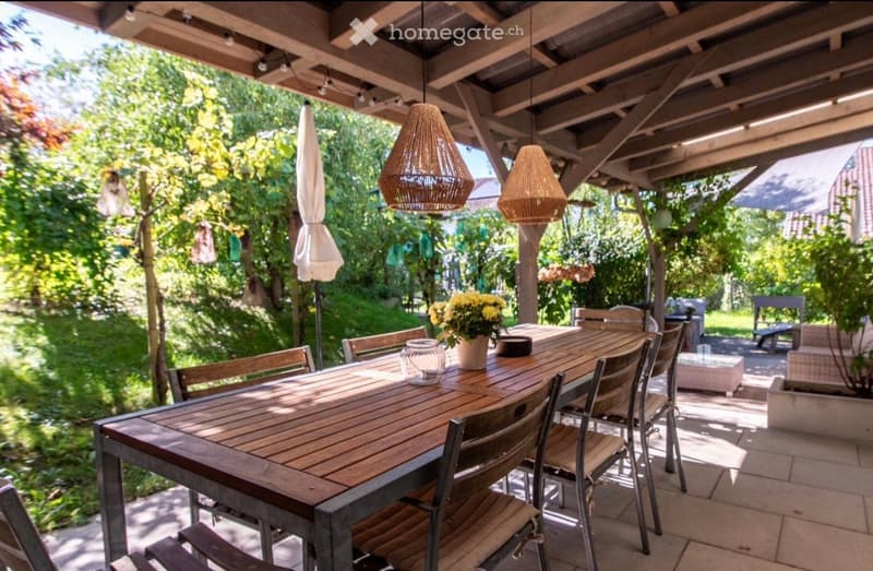 Wooden table with chairs, hanging lamps, flower vase, closed umbrella, paved floor, green plants, trees