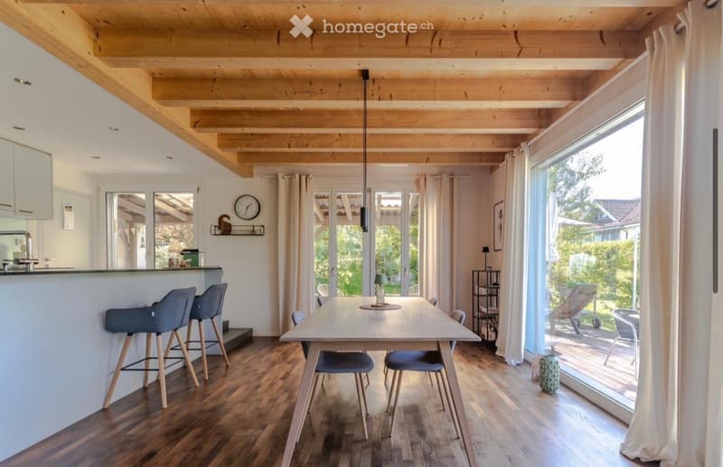 Dining room with wooden floor, wooden ceiling beams, large windows, table, chairs, kitchen counter, bar stools, clock, shelves, lamp, and a view of the garden