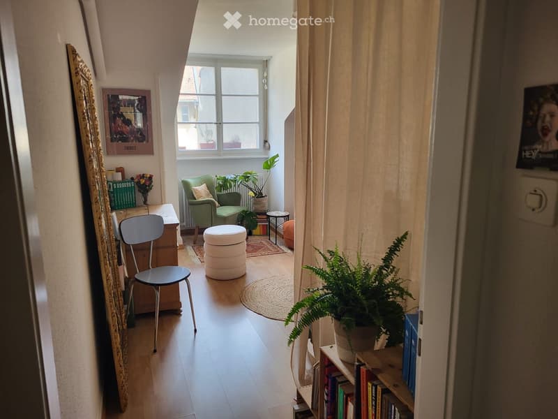 A room with wooden floor, a green chair, a white stool, a desk, a mirror, a plant, a window, and a curtain.