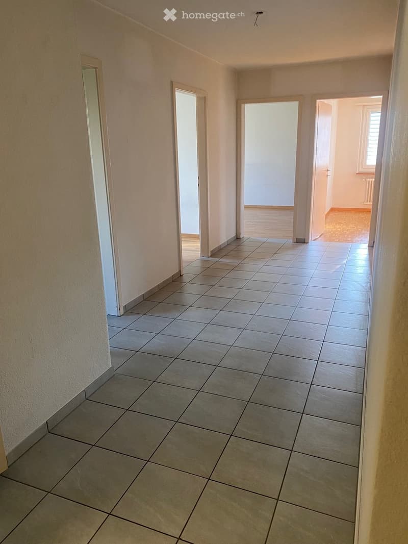 Empty hallway with tiled floor, three doors, one leading to a room with wooden floor and window, another leading to a room with radiator and window, and the last one leading to a room with wooden floor and window.