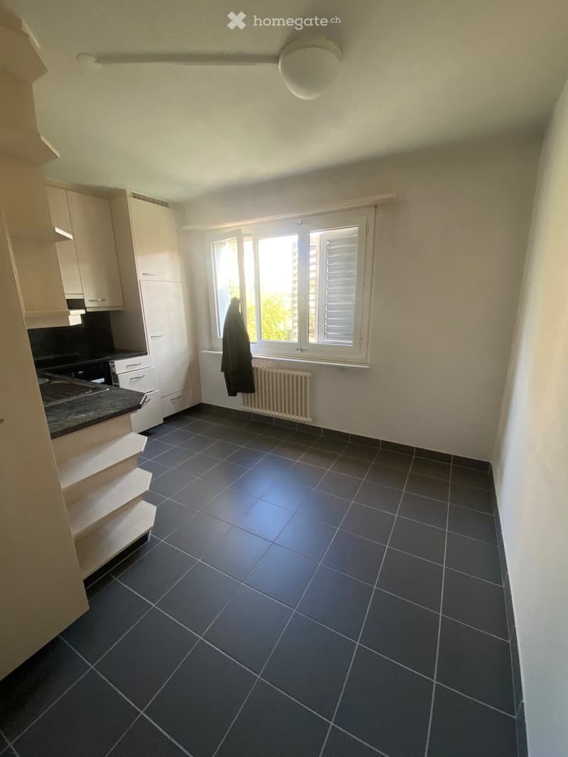 Empty kitchen with white cabinets, black tiled floor, white walls, window with white shutters, white ceiling light, white radiator