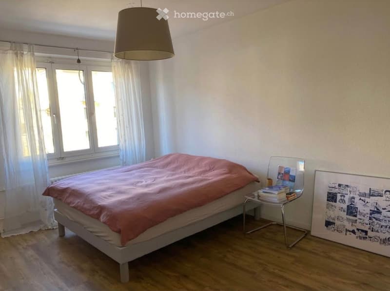 Single bed, white walls, wooden floor, white curtains, lamp, small table with books, picture frame