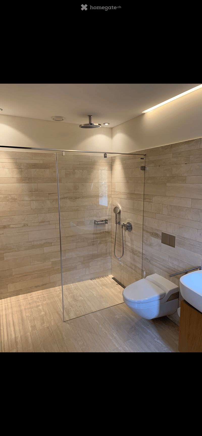 Modern bathroom with glass shower enclosure, beige tiles, white toilet, white sink, wooden cabinet, ceiling lights