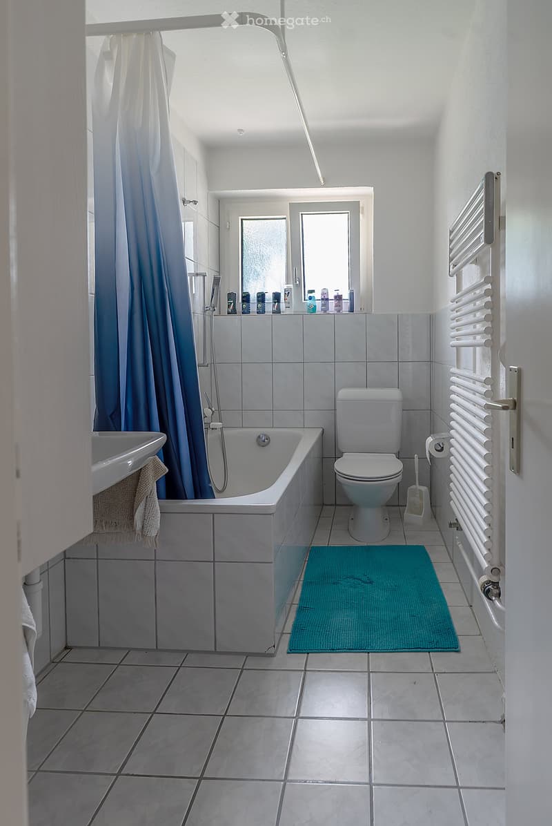 Bathroom with bathtub, toilet, sink, and towels. Blue rug on the floor and white tile walls. Window above the bathtub.