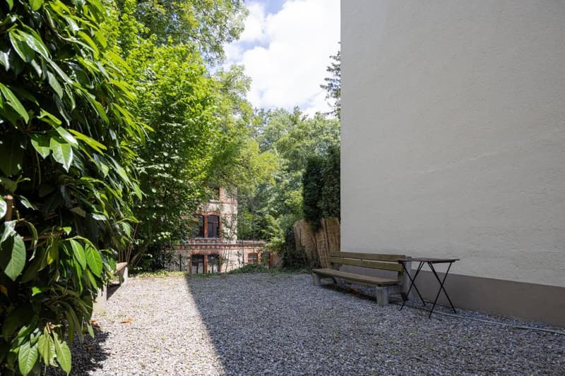 A gravel path leads to a wooden bench and a table. Behind the bench is a white wall. Trees and shrubs surround the area. The shadows of the trees fall on the ground. A building with brick walls is visible behind the plants.