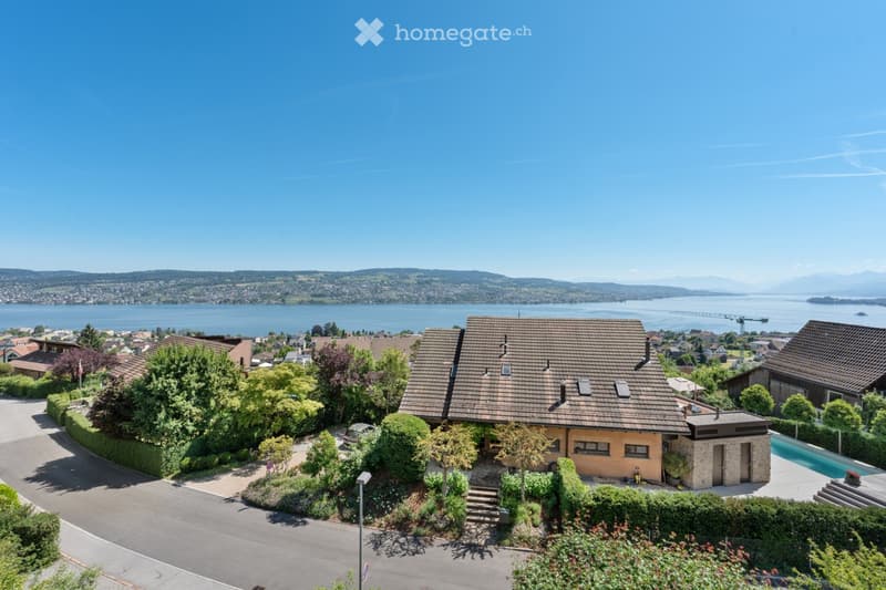 A two-story house with a tiled roof, surrounded by greenery and a swimming pool, overlooking a scenic lake and distant mountains.
