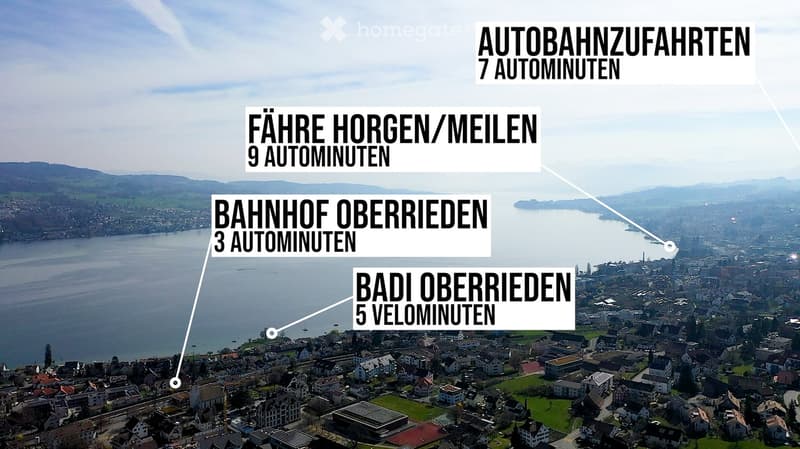 Aerial view of a town with a lake, mountains, and a railway station. The town has many buildings, some with solar panels, and a green field. The lake is calm, and the sky is clear. The text on the image provides information about the travel time to differ