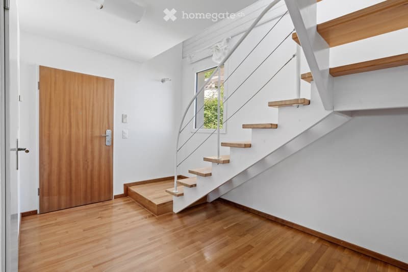 Modern hallway with wooden flooring, white walls, and a staircase featuring wooden steps and a white metal railing. A wooden door is visible on the left side.