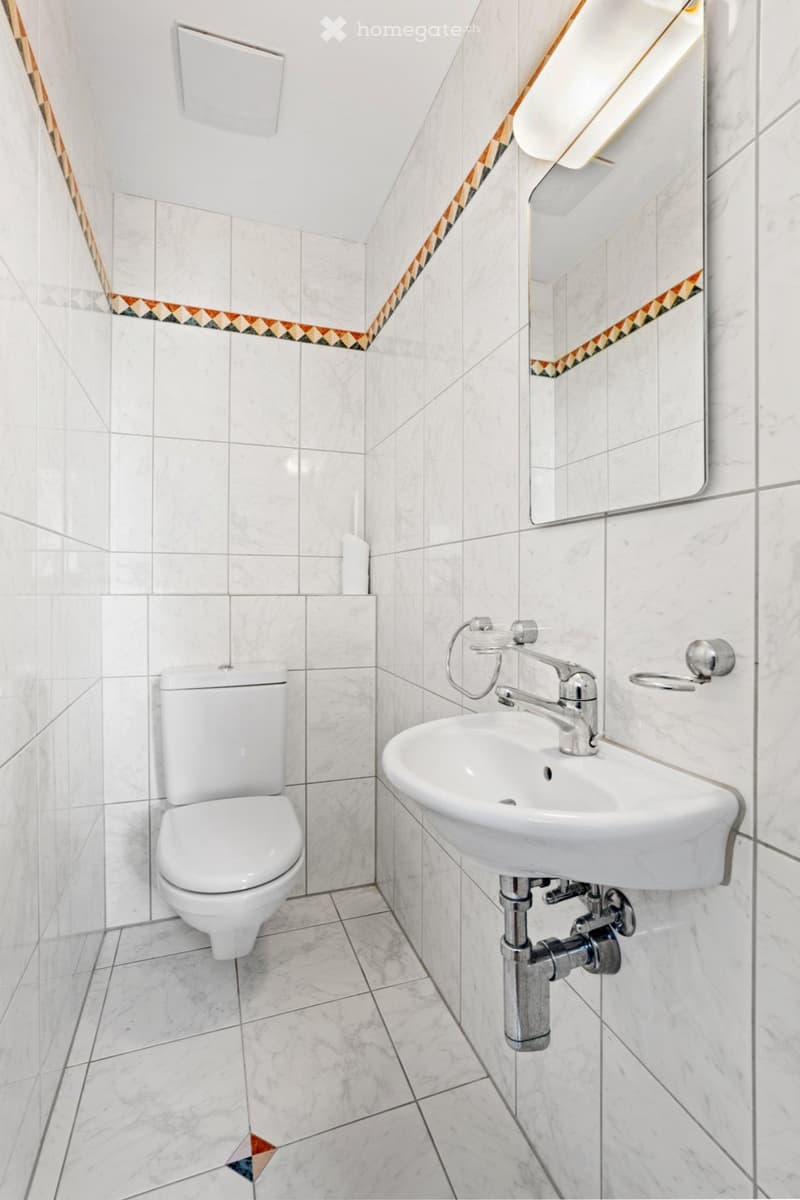 Modern bathroom with white tiles, a toilet, a sink with a chrome faucet, and a large mirror with a decorative border.
