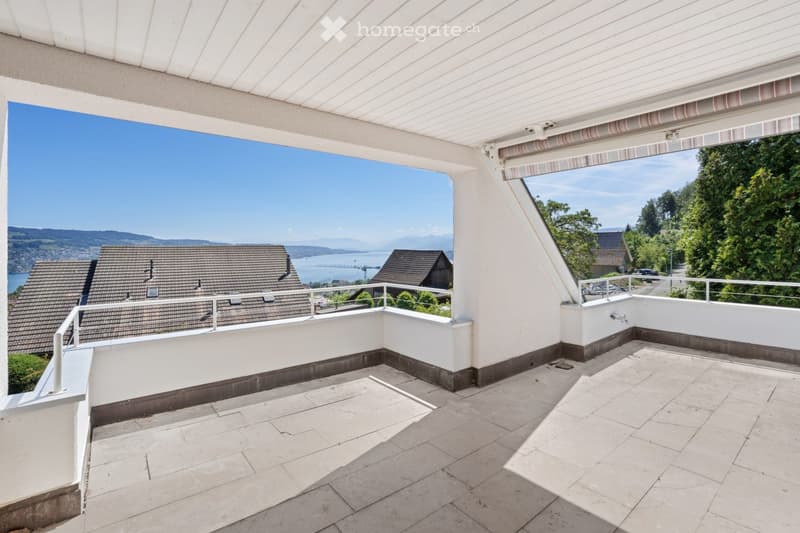 Balcony with a panoramic view of a lake and surrounding landscape, featuring a tiled floor, white walls, and a retractable awning.