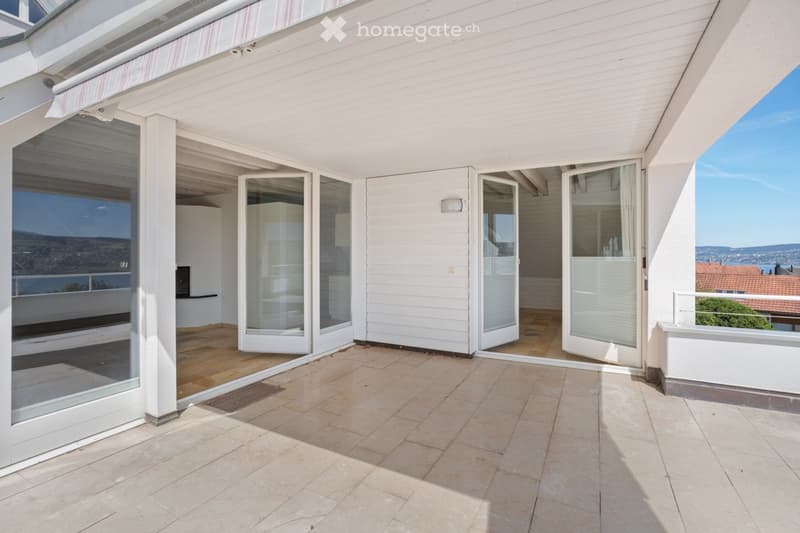 A spacious, modern balcony with large glass sliding doors leading to an indoor area, tiled flooring, and a view of the surrounding landscape.