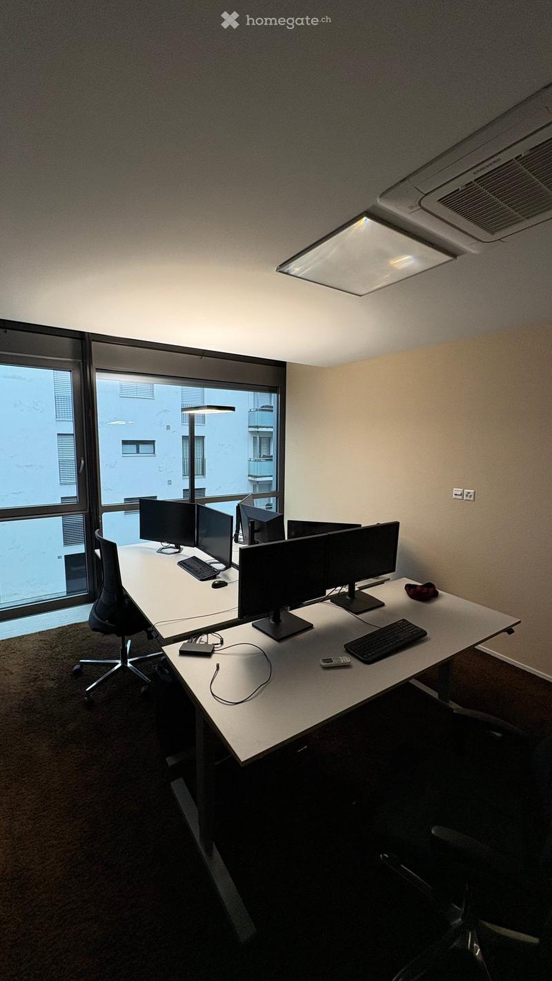 An office room with two desks, each with three monitors, a keyboard, a mouse, and a phone. The room has a carpeted floor, a glass window with a view of a building, and a ceiling with an air conditioner and a lamp.