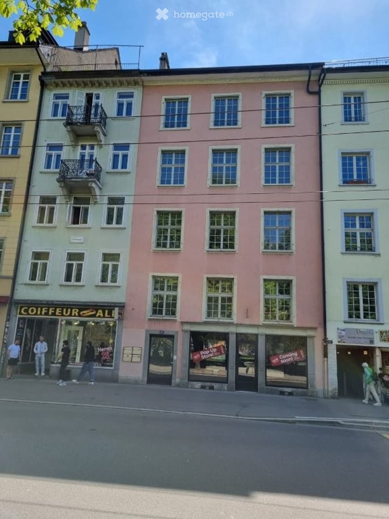 A multi-story building with multiple windows and balconies, a shop on the ground floor, and people walking on the sidewalk.