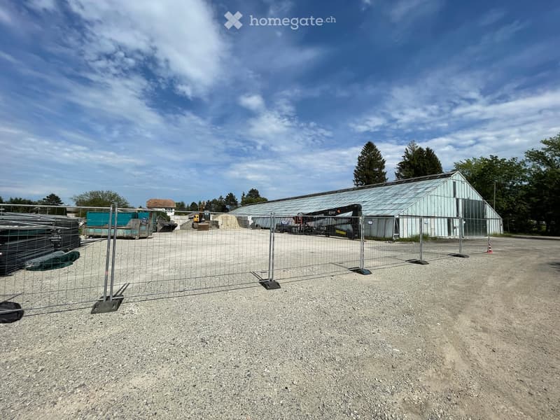 Industrial building with a metal roof, surrounded by a metal fence, with a machinery vehicle parked in front.