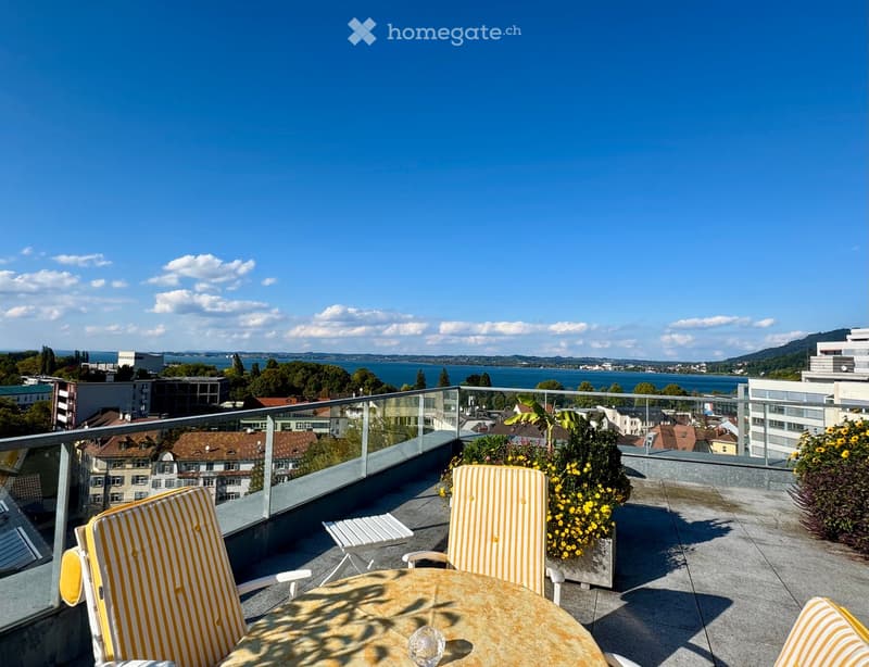 Terrace with table and chairs, yellow flowers, glass railing, view of the city and lake