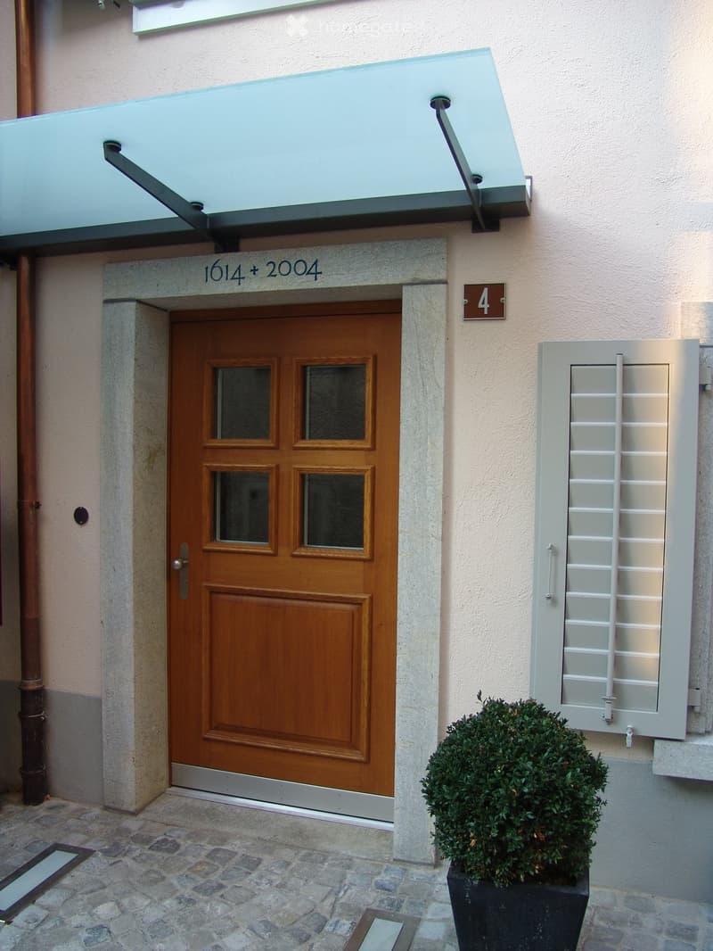 Exterior of a house with a wooden door, glass windows, a glass canopy, and a potted plant.