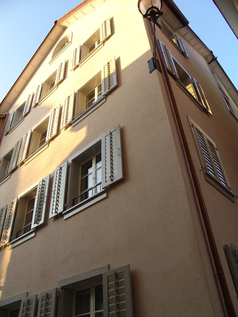A multi-story building with multiple windows and shutters, beige exterior, and a lamp on the wall.